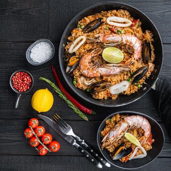 Typical spanish seafood paella in traditional pan and black bowl on black wooden background, flat lay, food photo.