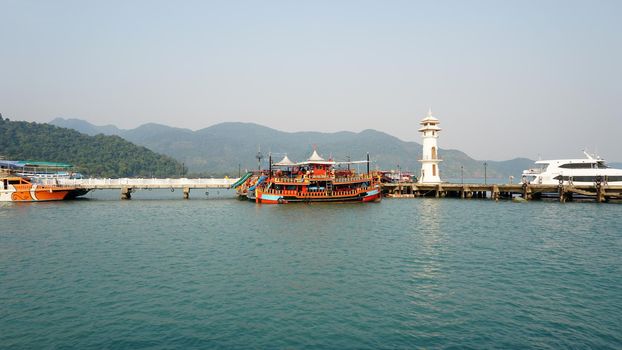 Sea port of Koh Chang, Thailand. There are ships. Green hills covered with haze, many trees and palm trees. Grey sky and emerald water. Pier in the sea. There are modern ships and Thai boats.