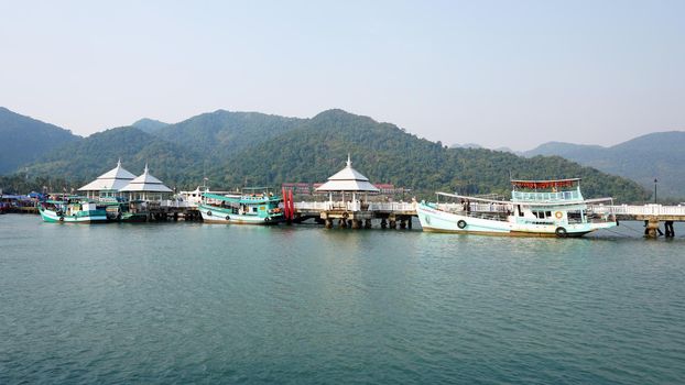 Sea port of Koh Chang, Thailand. There are ships. Green hills covered with haze, many trees and palm trees. Grey sky and emerald water. Pier in the sea. There are modern ships and Thai boats.