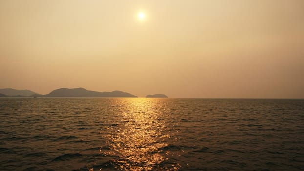 Sunset on an island with a sea view. A ray of sun is reflected in the water. The yellow-red sun is visible in the distance. People are resting. Hills of the island, palm trees and a light haze.