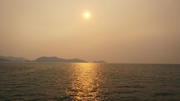 Sunset on an island with a sea view. A ray of sun is reflected in the water. The yellow-red sun is visible in the distance. People are resting. Hills of the island, palm trees and a light haze.