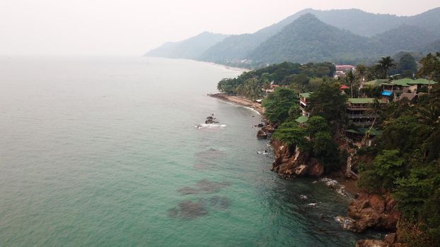 Green Chang island is covered in smoke from a fire in Cambodia. Green trees, palm trees and hills are covered with clouds of smog. Hotels stand on a cliff in front of the sea. Turquoise water.