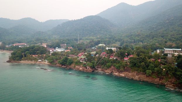 Green Chang island is covered in smoke from a fire in Cambodia. Green trees, palm trees and hills are covered with clouds of smog. Hotels stand on a cliff in front of the sea. Turquoise water.