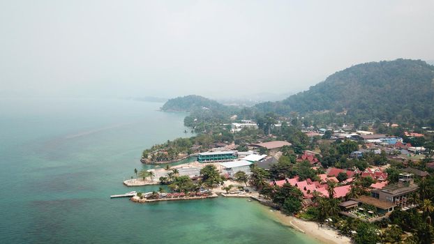 Green Chang island is covered in smoke from a fire in Cambodia. Green trees, palm trees and hills are covered with clouds of smog. Hotels stand on a cliff in front of the sea. Turquoise water.