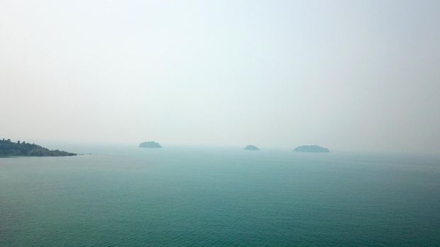 Green Chang island is covered in smoke from a fire in Cambodia. Green trees, palm trees and hills are covered with clouds of smog. Hotels stand on a cliff in front of the sea. Turquoise water.