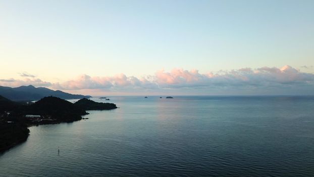 Dawn on Chang Island. Turquoise clear water with waves reflects the environment. Light clouds of pink color, green hills, palm trees. A sharp rocky slope into the water. Shooting from drone. Thailand
