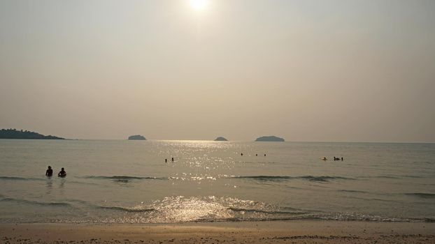 Smog obscures the sun's rays. People relax on the beach. Yellow rays of light. View of the islands, sand, sea and palm trees. Dirty air, smog. Chang Island, Thailand.