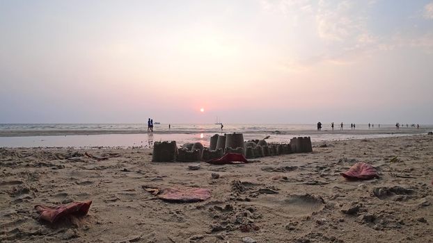 Sand castle on the beach. Sunset on the island. Purple leaves lie on the sand. Traces of people are visible. Families, children, and couples walk in the distance. Yachts are visible in sea. Thailand