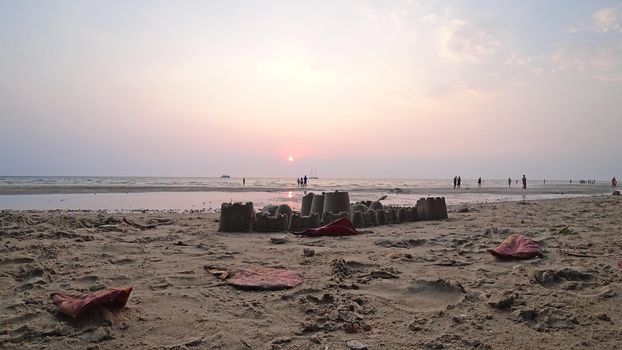 Sand castle on the beach. Sunset on the island. Purple leaves lie on the sand. Traces of people are visible. Families, children, and couples walk in the distance. Yachts are visible in sea. Thailand