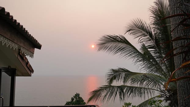 Sunset on an island with a sea view. A ray of sun is reflected in the water. The yellow-red sun is visible in the distance. People are resting. Hills of the island, palm trees and a light haze.