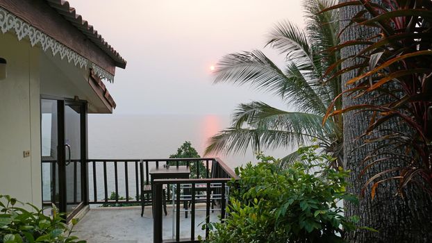 Sunset on an island with a sea view. A ray of sun is reflected in the water. The yellow-red sun is visible in the distance. People are resting. Hills of the island, palm trees and a light haze.