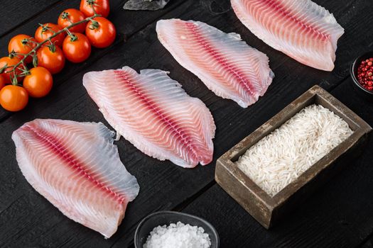 Raw white fish tilapia, with basmati rice and cherry tomatoes ingredients, on black wooden background
