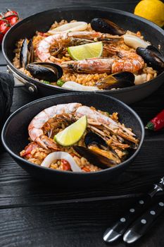 Typical spanish seafood paella in traditional pan and black bowl on black wooden background, food photo.
