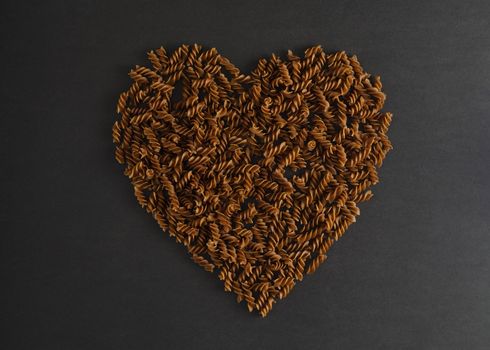 Minimalistic flat lay with heart made of buckwheat pasta fusilli on dark background. Concept of cooking healthy or vegetarian gluten free food