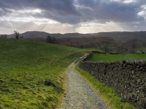 Path by a dry-stone wall in the countryside with rays of sunlight bursting through a dark and stormy sky, Lake District, UK