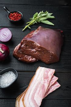 Raw beef liver with bacon and onion on black wooden background.
