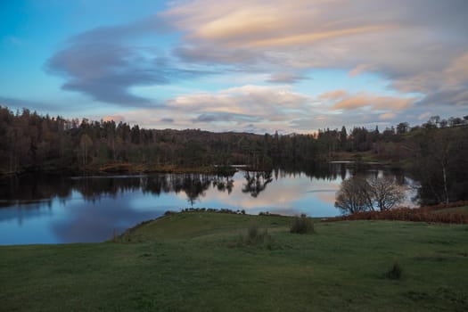View over the calmness of Tarn Hows at sunset with the fells in the background and colourful clouds reflected in the water, Lake District, UK