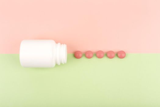 Top view of an opened white medication bottle with spilled red round pills on green and pink background. Concept of pharmacy and medications