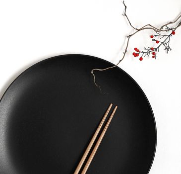 Black round plate, two chopsticks and Japanese bamboo in soft focus on white background. Japanese dining inspiration, table arrangement