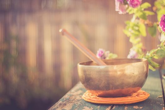 Metal singing bowl on a rustic green, wooden table outdoors. Flowers in the colourful, blurry background