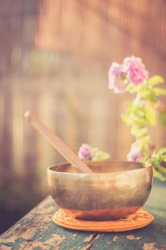 Metal singing bowl on a rustic green, wooden table outdoors. Flowers in the colourful, blurry background