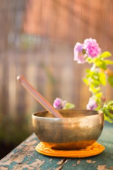 Metal singing bowl on a rustic green, wooden table outdoors. Flowers in the colourful, blurry background