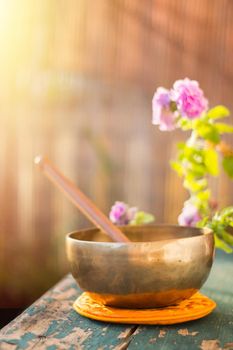 Metal singing bowl on a rustic green, wooden table outdoors. Flowers in the colourful, blurry background