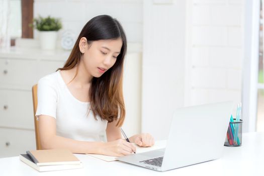 Young asian woman writing on notebook for planning working and using laptop computer on desk at home, girl notes about finance, female study and learning, business and communication concept.