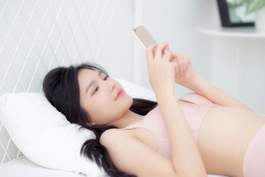 Beautiful young asian woman sexy in underwear chatting on mobile phone in the bedroom, happy girl in lingerie looking social media on smartphone lying on bed, communication and lifestyle concept.