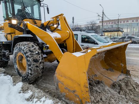 TULA, RUSSIA - NOVEMBER 21, 2020: Yellow tractor with large scoop cleaning snow on road at winter day light.