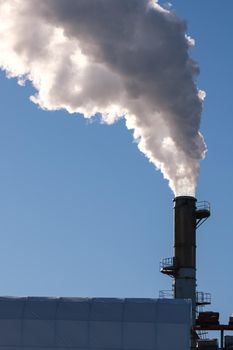 Thick smoke billows into a blue sky from a tall smokestack in a vertically-oriented image.