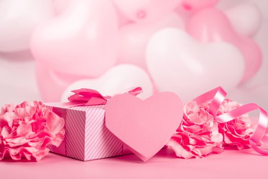 Valentine Day gift in a box wrapped in striped paper and tied with silk ribbon bow and heart shaped greeting card on pink balloons background with copy space for text
