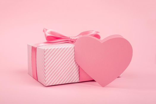 Valentine Day gift in a box wrapped in striped paper and tied with silk ribbon bow and heart shapes greeting card on pink background with copy space for text