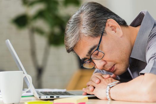 Asian businessman tired overworked he sleeping over a laptop computer on the desk. senior man with eyeglasses lying asleep on table at his working place