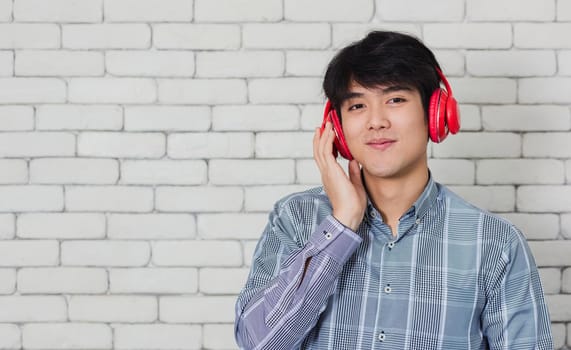 Portrait of Asian handsome young man he smile listening to music in headphones on brick wall background. Happy teenage guy smiling. Happy laughing man standing with red headphones