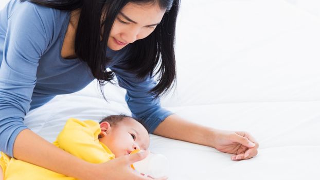Portrait of beautiful young Asian mother holding and feeding infant newborn baby from milk bottle in a white bed, The child drinking milk from mom, Healthcare and medical