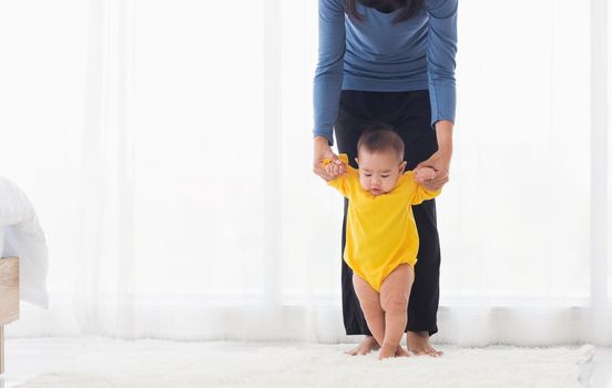 Asian little baby girl taking first steps learning to walk with mom help support the cute unstable walking toddler at home in bedroom. Happy family first steps parenthood concept