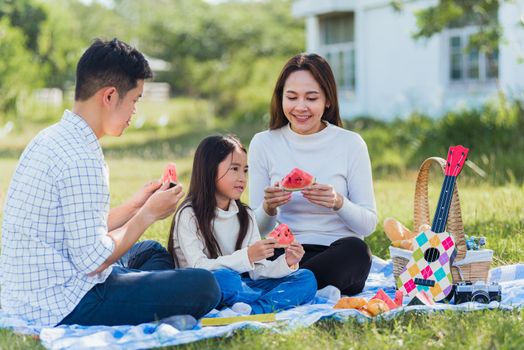 Happy Asian young family father, mother and child little girl having fun and enjoying outdoor sitting on picnic blanket eating watermelon fruit snack lunch in park sunny time, summer leisure concept
