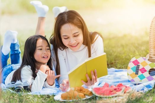 Happy Asian young family mother and child little girl having fun and enjoying outdoor laying on picnic blanket reading book at summer garden spring park, Family relaxation concept