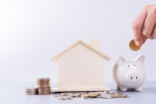 Hand putting money coin into piggy bank with wooden house and stack coins isolated grey background. Property investment and house mortgage financial concept.