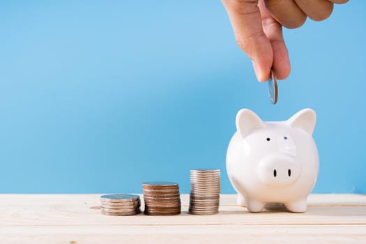 Hand putting money coin into piggy bank with stack coins isolated blue background. Saving money and investment concept