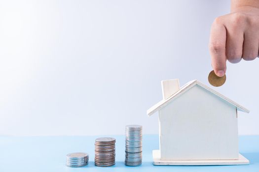 Hand putting money coin into wooden house with stack coins isolated grey background. Property investment and house mortgage financial concept.