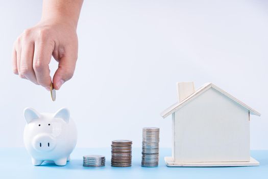 Hand putting money coin into piggy bank with wooden house and stack coins isolated grey background. Property investment and house mortgage financial concept.