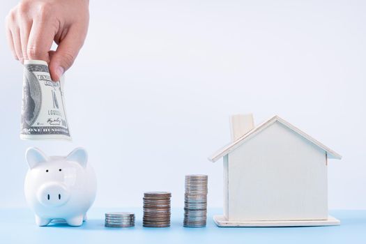 Hand putting money paper into piggy bank with wooden house and stack coins isolated grey background. Property investment and house mortgage financial concept.