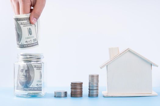 Hand putting money paper into jar with wooden house and stack coins isolated grey background. Property investment and house mortgage financial concept.