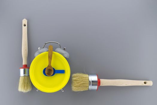 Creative DIY, decorating or renovations background with two clean bristle brushes and a colorful yellow alarm clock with tools for hands forming a border on grey with copy space