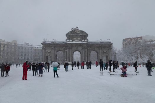 Madrid, Spain - January 09, 2021: The famous Alcala gate, frozen and covered with snow, surrounded by people walking, on a snowy day, due to the polar cold front Filomena.