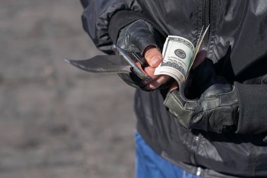close up, the hands of a bandit in leather gloves holding a knife and counting banknotes, dollars on the street