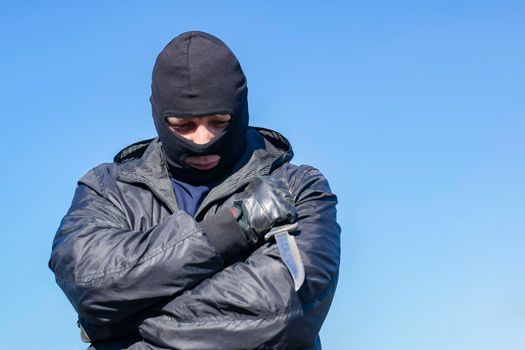 a sad, thoughtful bandit stands in a black mask and with a knife against the blue sky