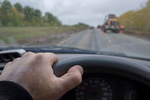 the driver's hand on the steering wheel of a car passing a road section under repair with a tractor, construction equipment in rainy cloudy weather